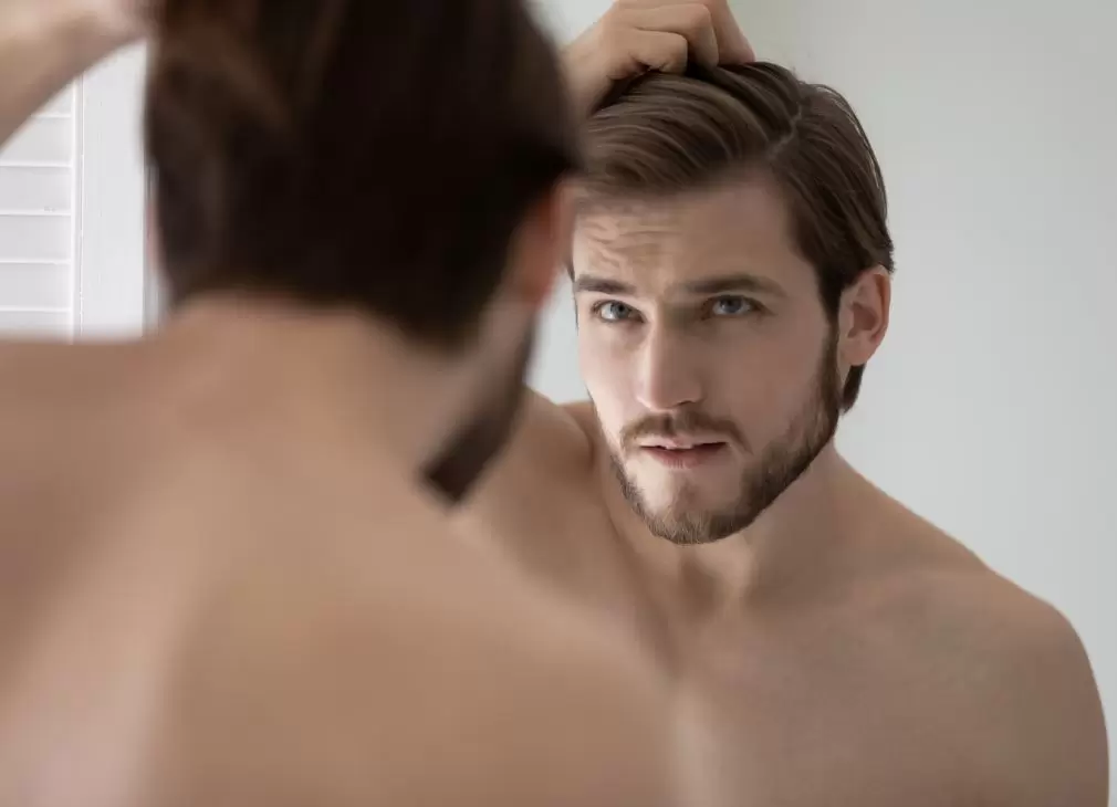 A young man with styled brown hair examines his reflection in a mirror while running his hand through his hair. He has a thoughtful expression, showcasing a well-groomed appearance. The setting is minimalistic, emphasizing his focus on personal grooming. This image captures themes of self-care and confidence, appealing to audiences interested in men's grooming and lifestyle.