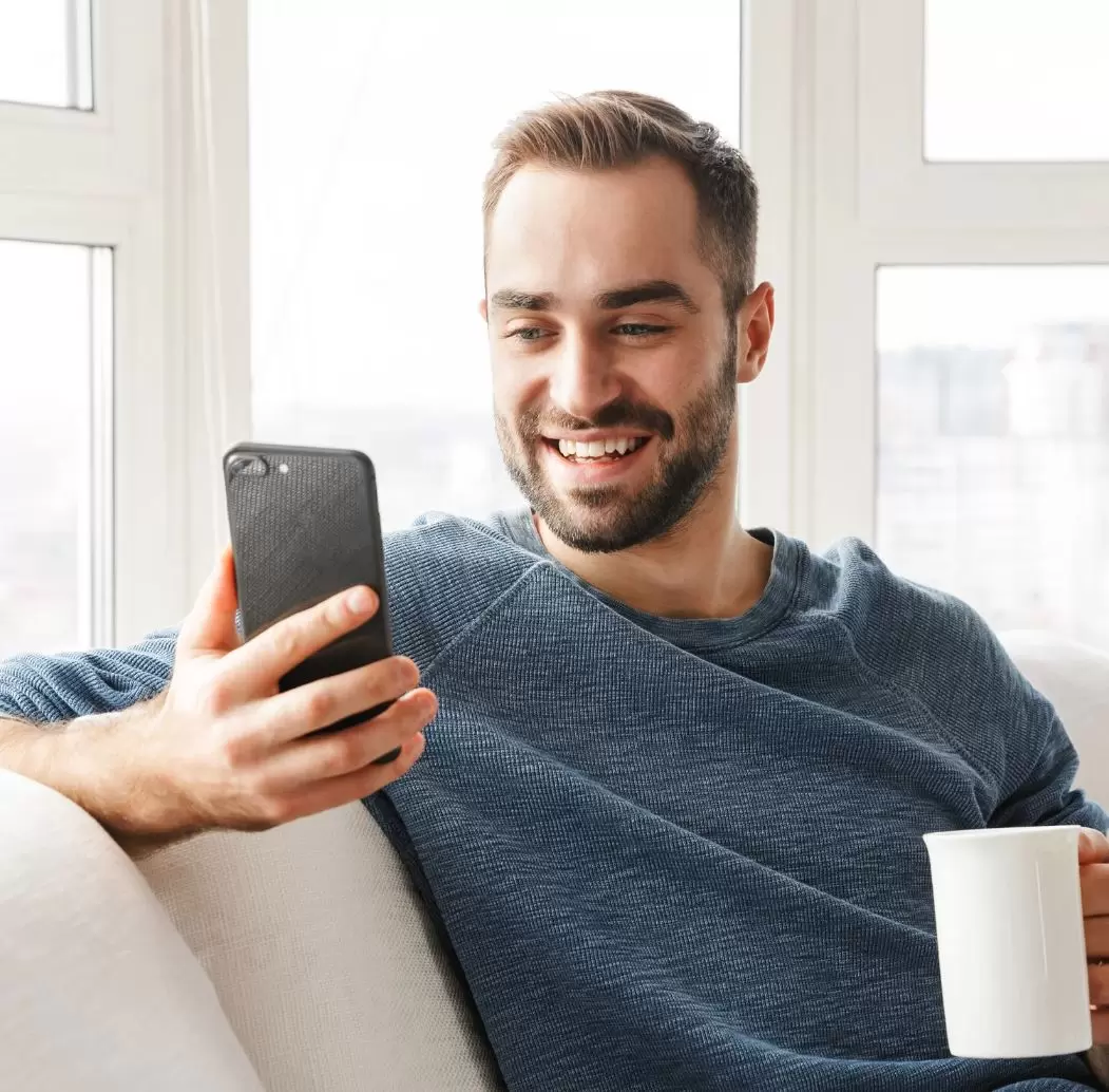 A man with a beard smiles while using a smartphone in a bright, cozy living room. He is seated on a sofa and holding a white coffee cup. Natural light streams through large windows, creating a warm atmosphere. The image conveys a sense of relaxation and enjoyment in a home setting, highlighting the connection between technology and everyday life. Ideal for articles on digital communication, work-from-home culture, or lifestyle trends.