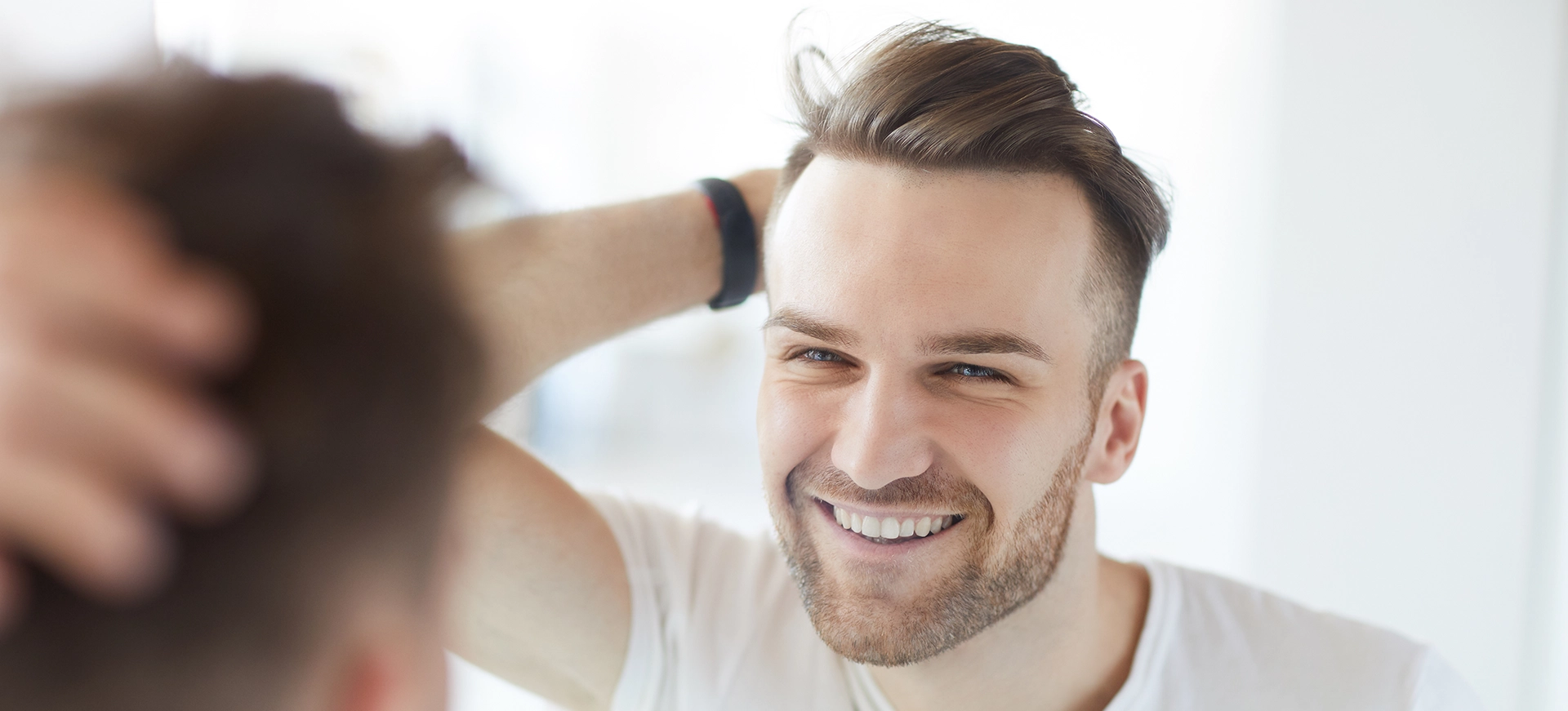 A young man with short, styled hair smiles at his reflection in a bright, modern bathroom. He is wearing a white t-shirt and is running his hand through his hair, showcasing a confident and relaxed demeanor. The background is softly blurred, emphasizing his expression and grooming routine. This image captures themes of self-care and personal grooming, appealing to those interested in hair styling and men's fashion.