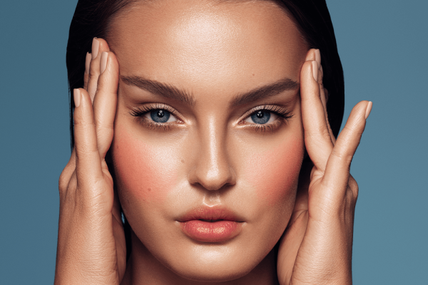 Close-up portrait of a young woman with flawless skin, striking blue eyes, and natural makeup. She gently holds her face with both hands, showcasing a serene expression. The background is a soft blue, enhancing her features. This image highlights beauty and skincare, making it ideal for topics related to cosmetics, facial care routines, and wellness.