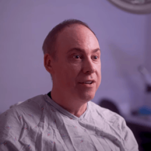 A man wearing a medical gown sits in a well-lit clinical setting, smiling and engaging in conversation. The background features medical equipment, indicating a healthcare environment. This image conveys a sense of comfort and professionalism, highlighting the importance of patient care in medical settings.