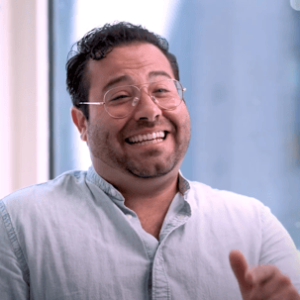 Smiling man with curly hair and glasses, wearing a light blue shirt, sitting indoors. He gestures playfully while engaging in conversation, conveying a friendly and approachable demeanor. The background features soft natural light, enhancing the welcoming atmosphere of the setting. This image captures a moment of joy and connection, ideal for content related to communication, teamwork, or positive interactions in a professional environment.